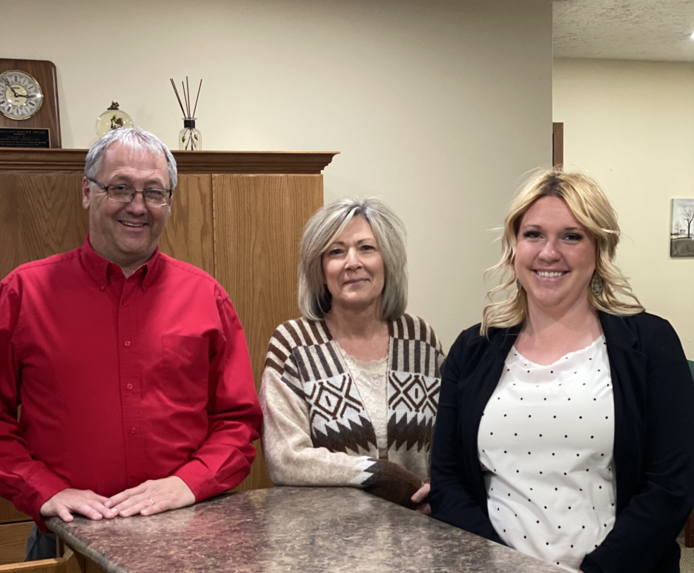 Three people sit behind a table in an office room, smiling at the camera.